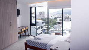 Quarto com uma janela grande, uma cama, uma escrivaninha e uma cadeira.