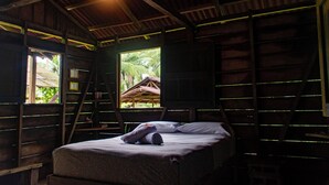 Una cama individual en una habitación con paredes de madera y techo inclinado. Se ve una ventana con vista a una palmera y un edificio.