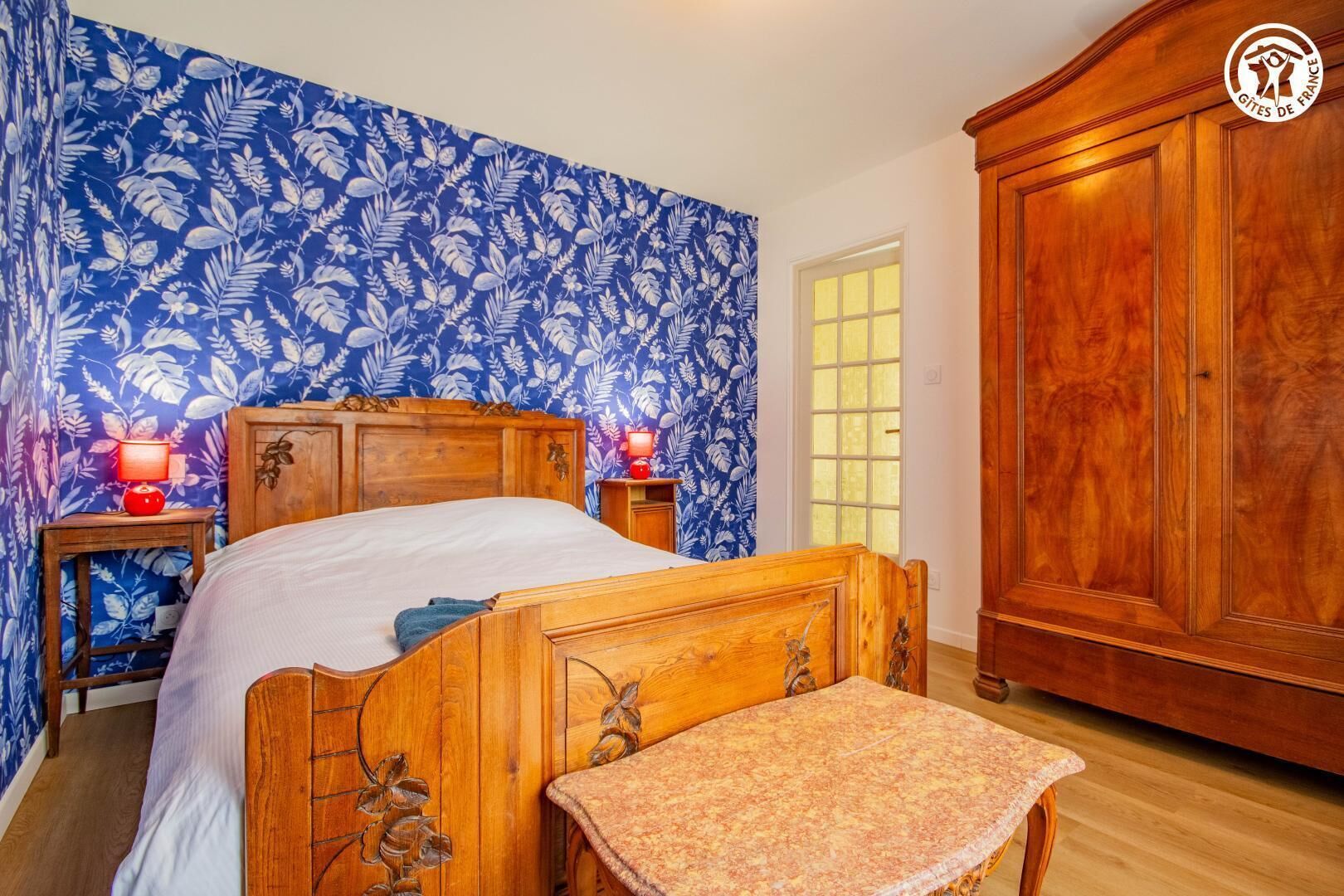 Une chambre à coucher avec un lit en bois, une table de chevet surmontée d’une lampe rouge, une armoire en bois et un mur orné d’un motif floral.