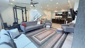 Ein modernes Wohnzimmer mit einem grauen Sofa, einem Hocker und einem gemusterten Teppich. Eine Deckenleuchte und Einbauleuchten sind sichtbar.