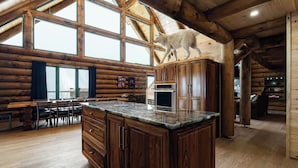 Interior de una cabaña de troncos con una gran mesa de comedor de madera, sillas, una isla de cocina y un lobo blanco en el techo.