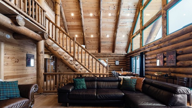 Interior de una cabaña de troncos con escalera de madera, sofás de cuero y una mesa de comedor de madera.