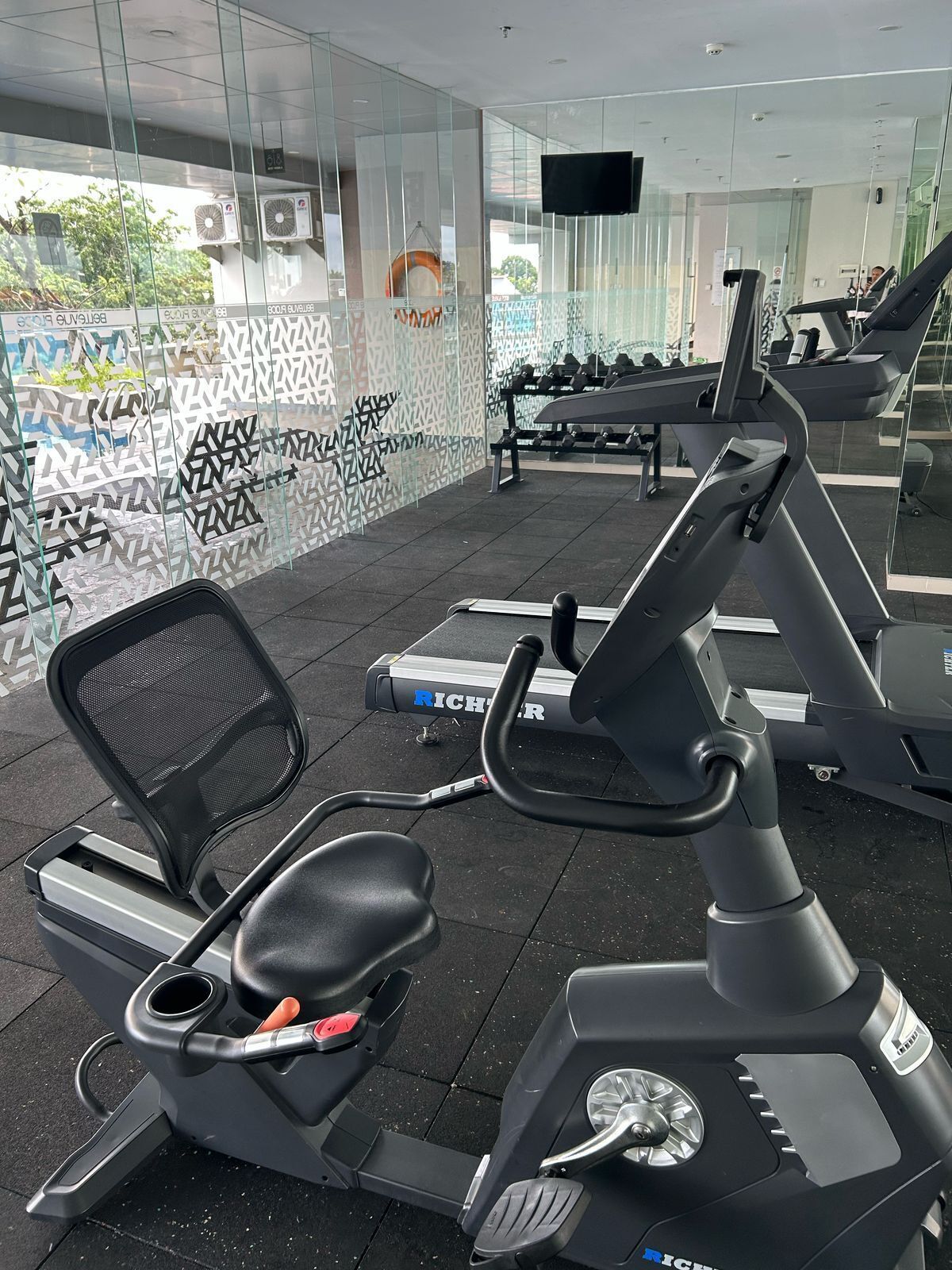 A modern gym with exercise equipment, a glass wall with a geometric pattern, and a television mounted on the wall.