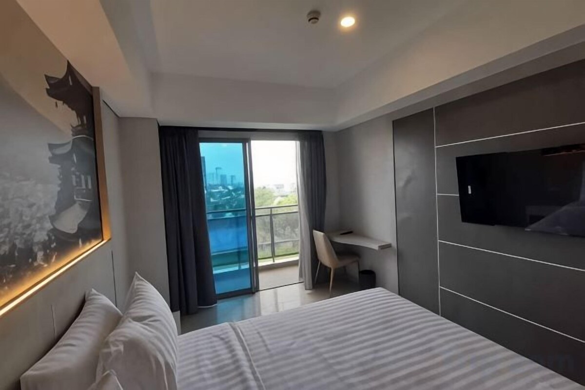 A modern hotel room with a large bed, a desk, a TV, and a balcony with a city view.