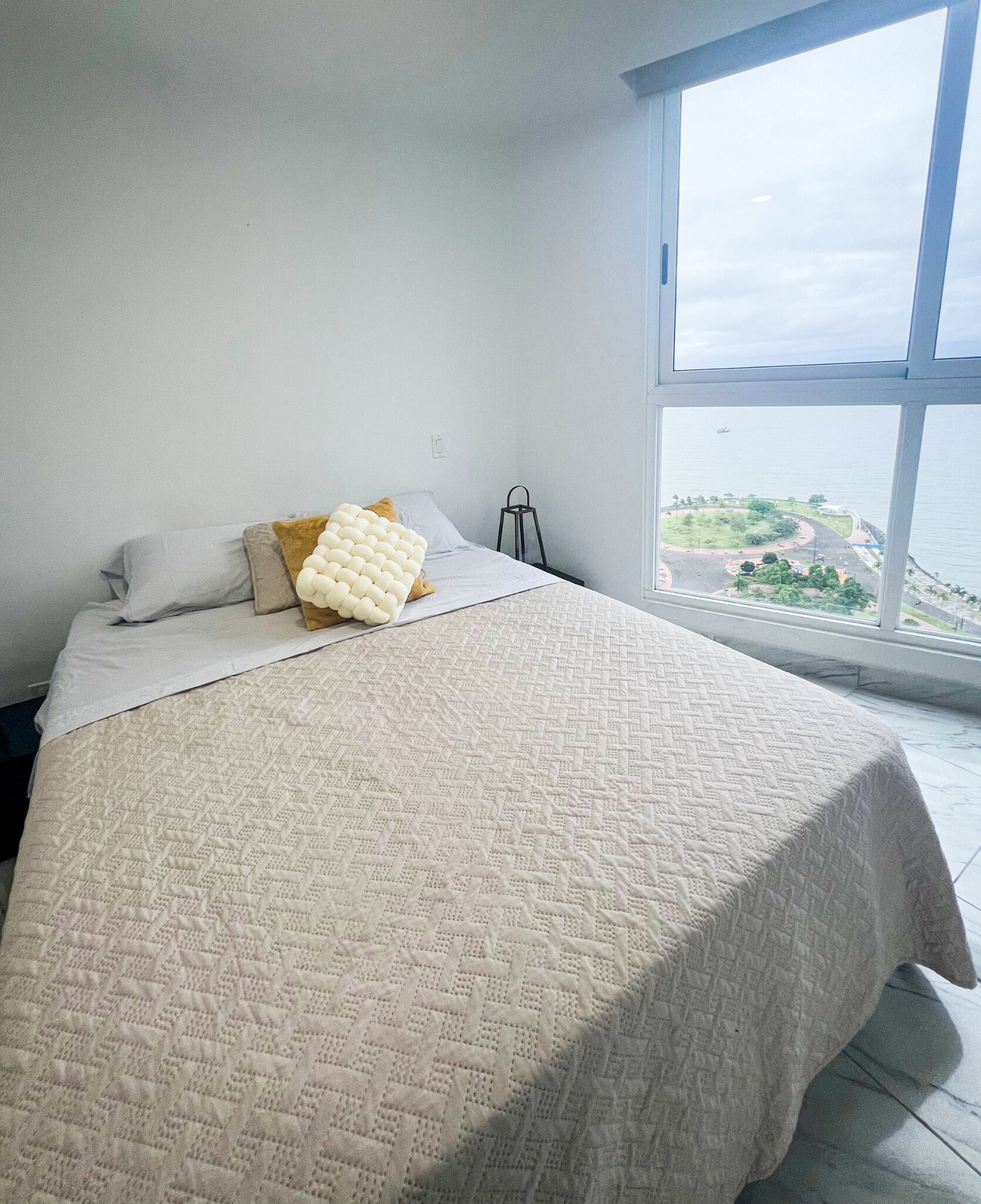 A bedroom with a large bed, a window with a view of a park and water, and a simple nightstand.