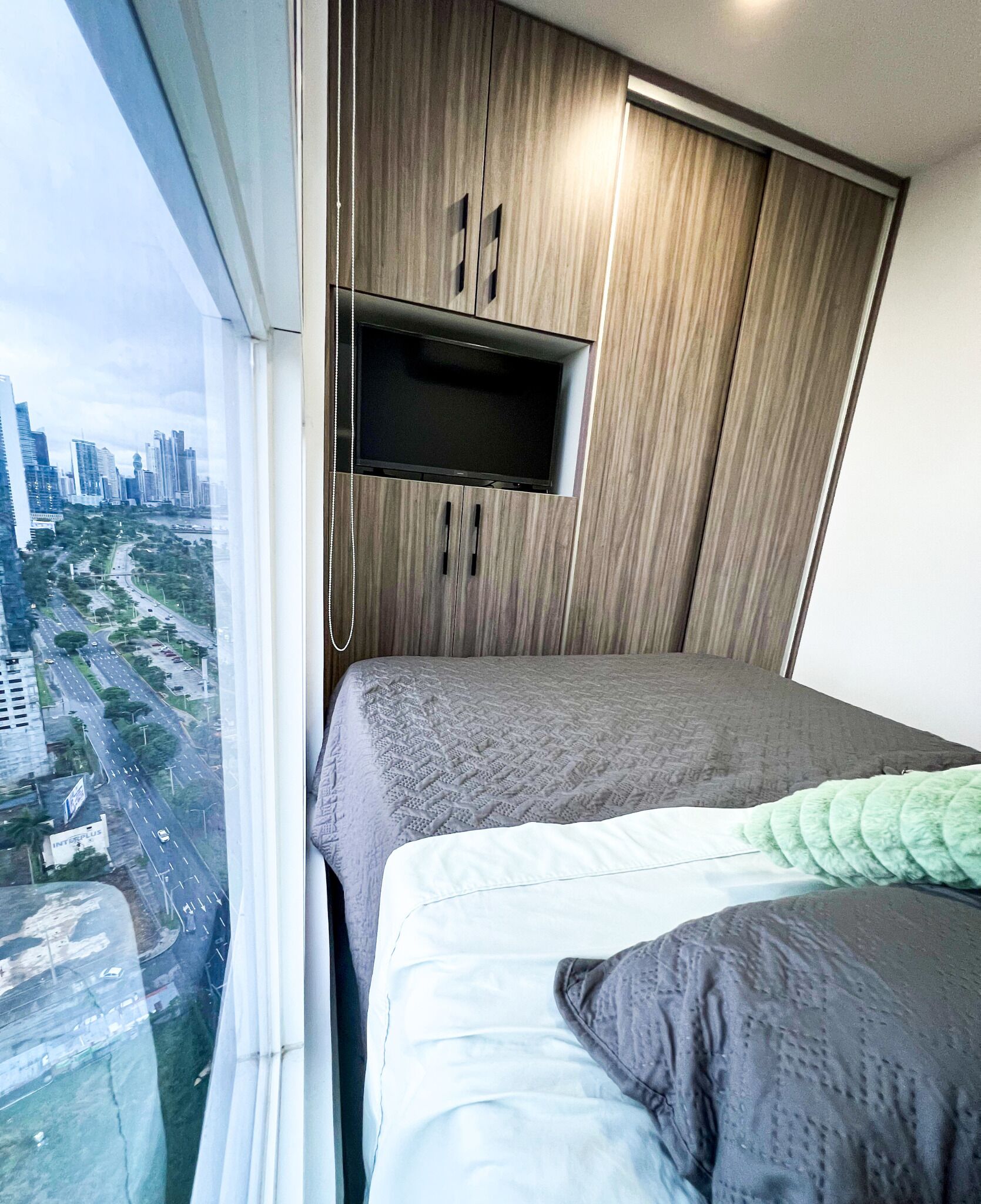 A bedroom with a city view, a bed with a grey and white duvet, a green throw pillow, and a TV mounted on a wooden cabinet.
