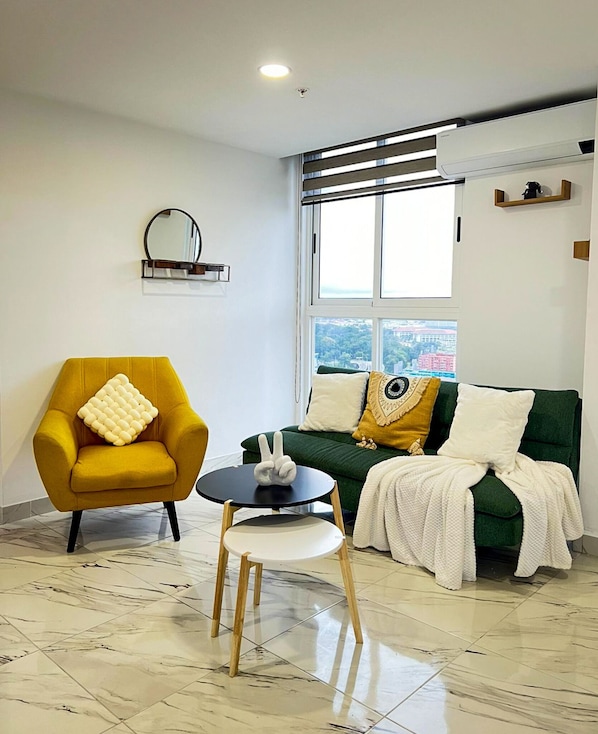 Un salon moderne avec un fauteuil jaune, un canapé vert agrémenté de plusieurs coussins, une petite table ronde et une grande fenêtre offrant une vue sur la ville.