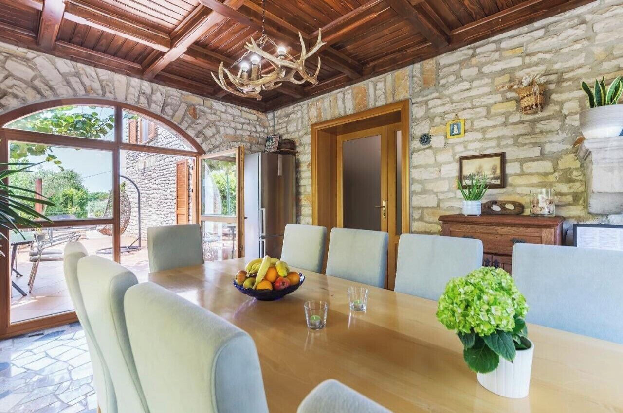 A dining area with a stone wall, wooden ceiling, and a chandelier. There is a table with chairs and a bowl of fruit.