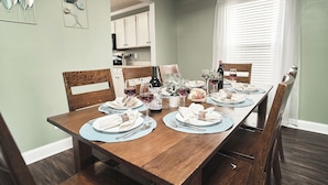 Une table à manger est dressée avec des assiettes, des verres et une bouteille, entourée de chaises en bois dans une pièce aux murs vert clair et dotée d’une fenêtre avec des rideaux blancs.