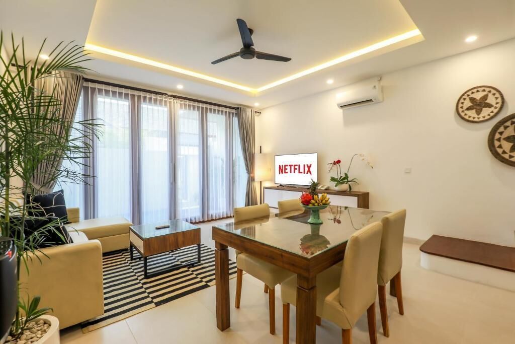 A modern living room with a glass dining table, beige sofas, a ceiling fan, and a TV displaying the Netflix logo.