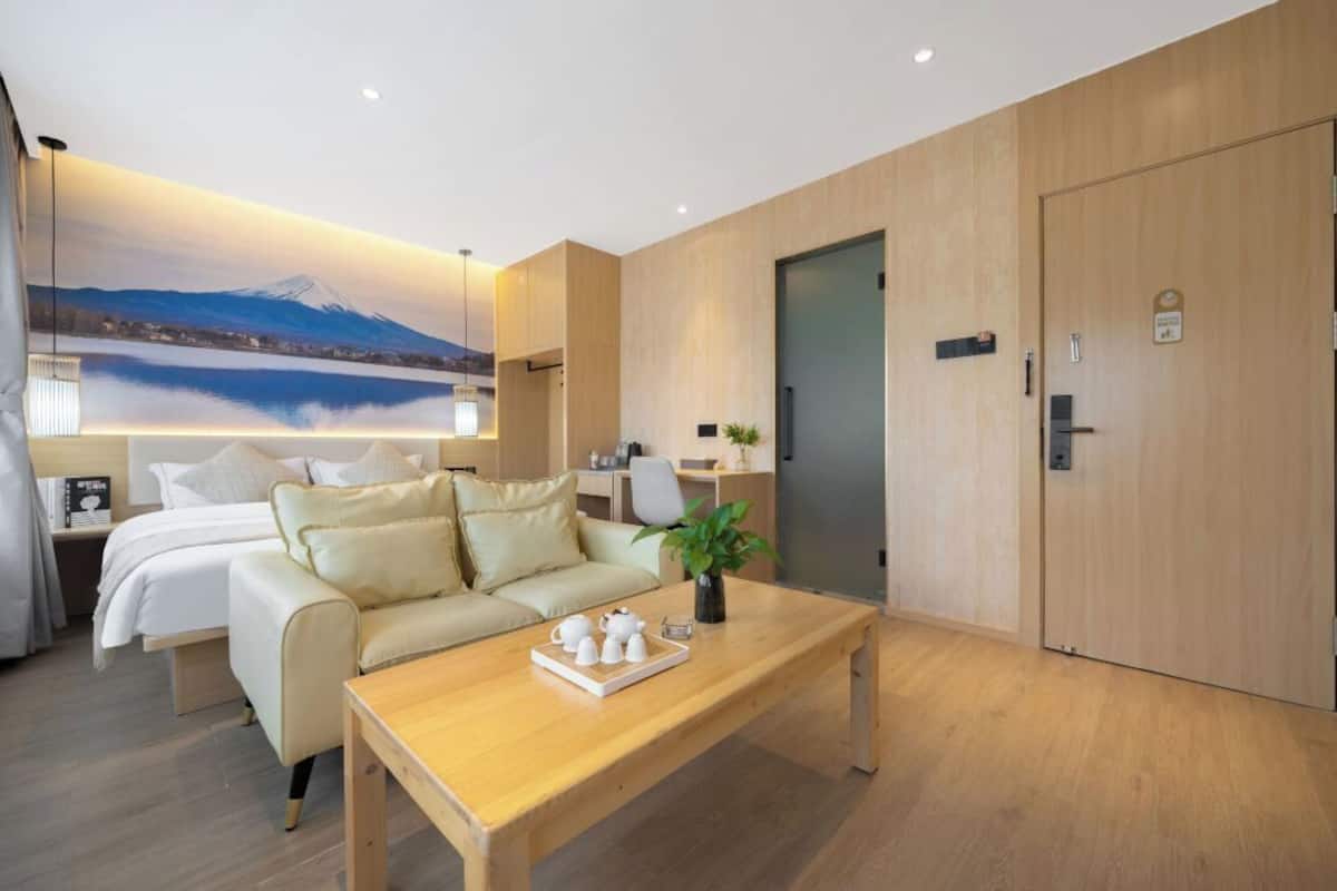 A modern hotel room with a bed, a sofa, a wooden coffee table, and a painting of a mountain landscape.