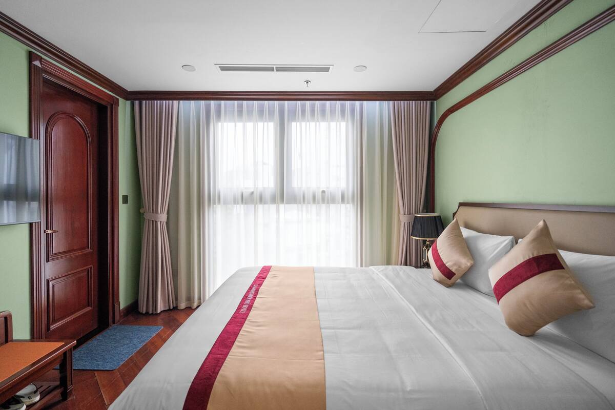 Habitación de hotel con una cama grande, una puerta de madera, un escritorio y una ventana con cortinas.