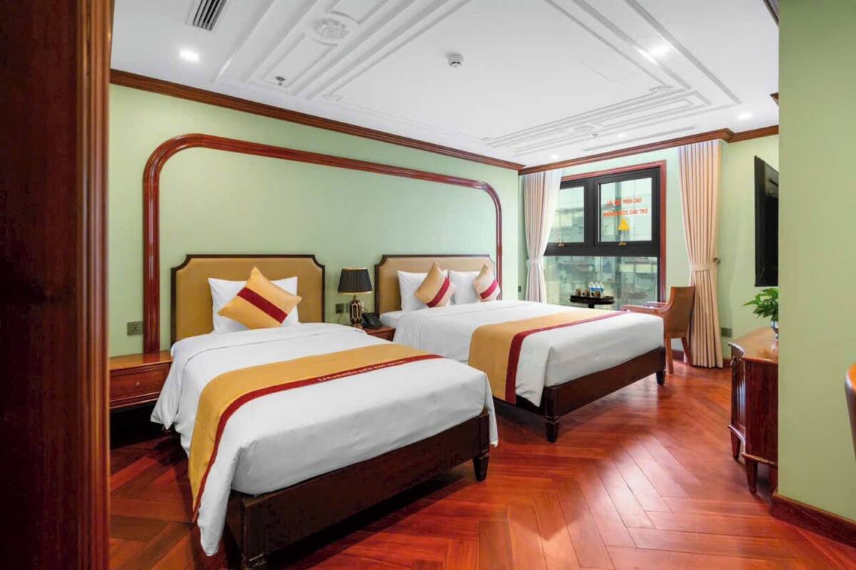Habitación de hotel con dos camas, piso de madera, un ventanal grande con cortinas y un televisor.