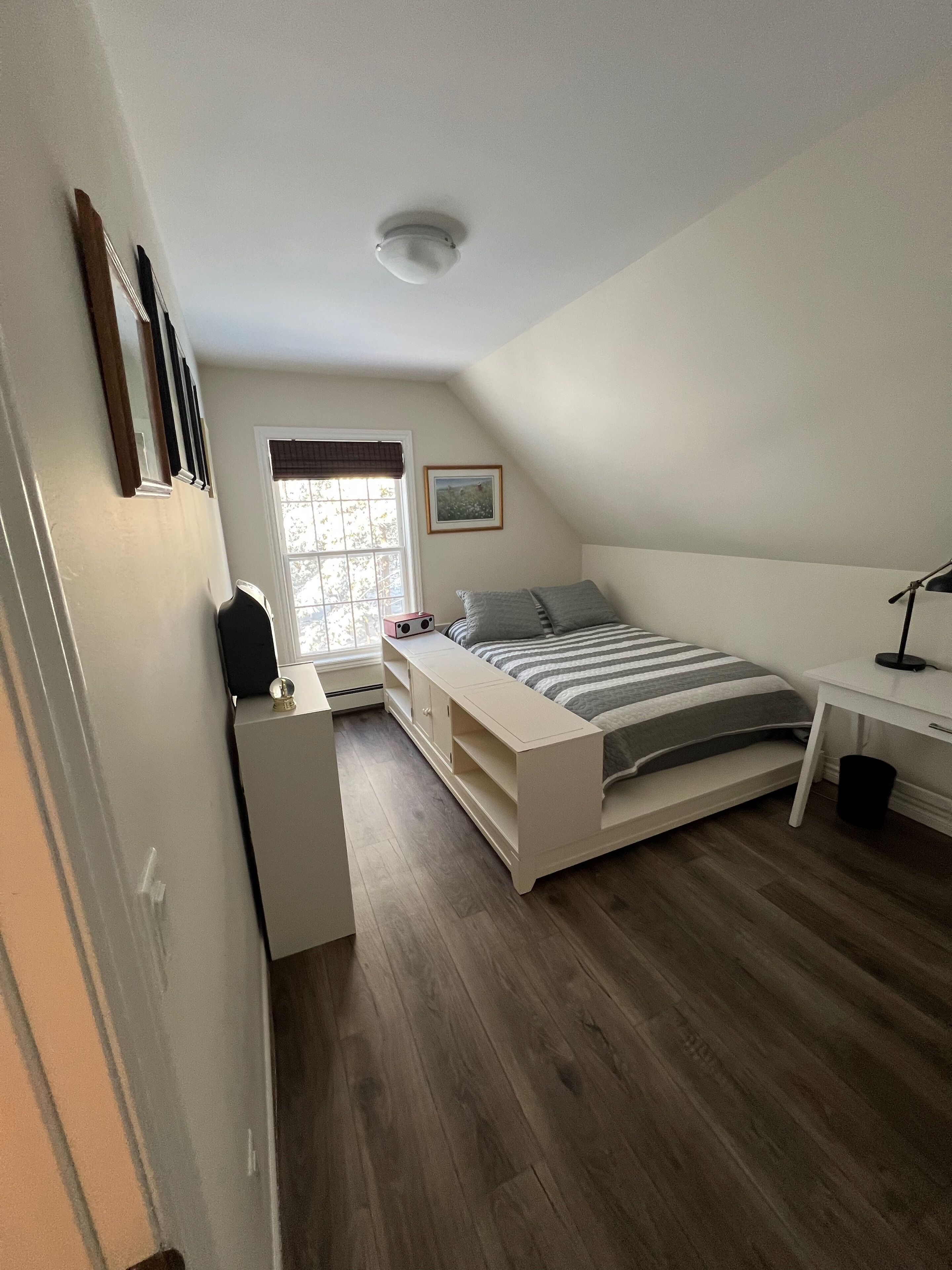 Una camera da letto con un letto, una scrivania, una finestra con persiane, una luce a soffitto e un quadro appeso al muro.