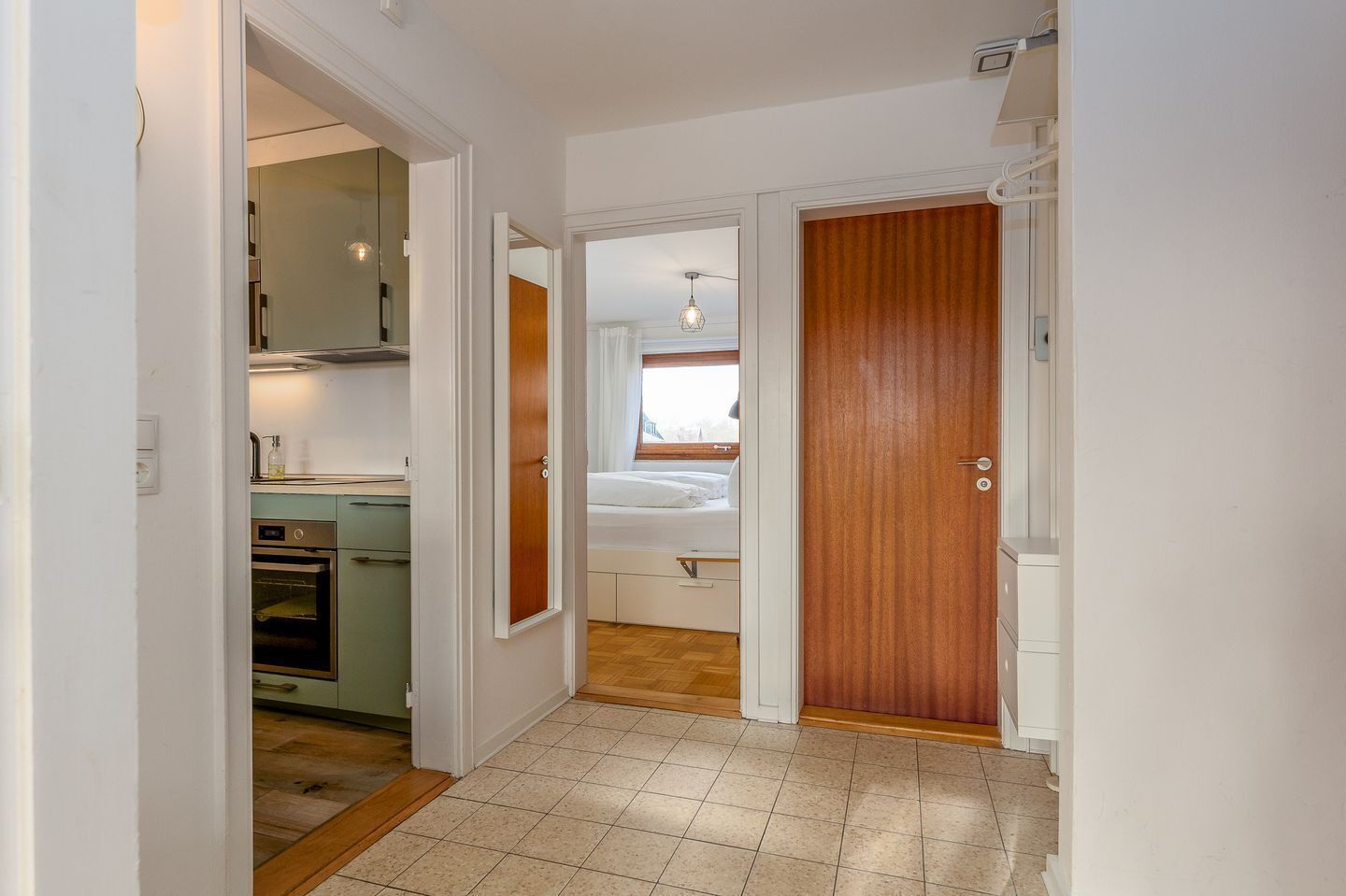 Een smalle gang met een keuken aan de linkerkant, een slaapkamer met een bed en raam, en een houten deur aan de rechterkant.