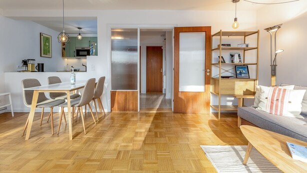 Ein modernes Wohnzimmer mit einer Couch, einem Couchtisch und einem Bücherregal. Es gibt einen Essbereich mit Tisch und Stühlen. Der Raum hat Holzböden und ein großes Fenster.