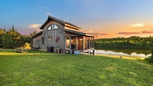 A rustic cabin with a porch, situated next to a lake, surrounded by lush greenery. - Cabin on Private 4-Acre Pond, King Bed (Junction City)