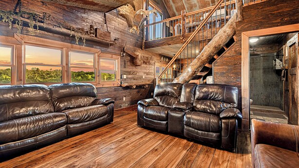 A rustic interior with wooden floors, leather sofas, a staircase, and large windows offering a view of the outdoors. - Cabin on Private 4-Acre Pond, King Bed (Junction City)