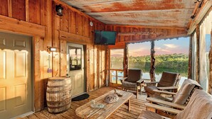 A wooden cabin with a TV mounted on the wall, a wooden table with a cushion, and a view of a lake through large windows. - Cabin on Private 4-Acre Pond, King Bed (Junction City)