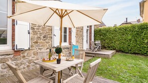 An outdoor wooden table with a beige umbrella, surrounded by wicker chairs, set against a stone wall with white shutters.