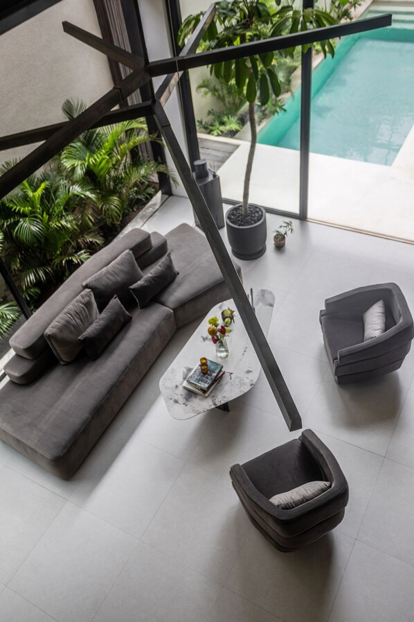 A modern living room with a grey sofa, two armchairs, a coffee table, and a view of a swimming pool through large windows. - Villa Sabilla Bingin (Pecatu)