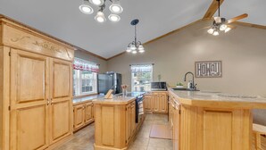 Une cuisine avec des meubles de rangement en bois, un îlot central, un ventilateur de plafond et une fenêtre avec des rideaux.