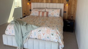 Una camera da letto con soffitto spiovente, pareti rivestite in legno, un letto con lenzuola decorate, due comodini con lampade e un quadro appeso al muro.