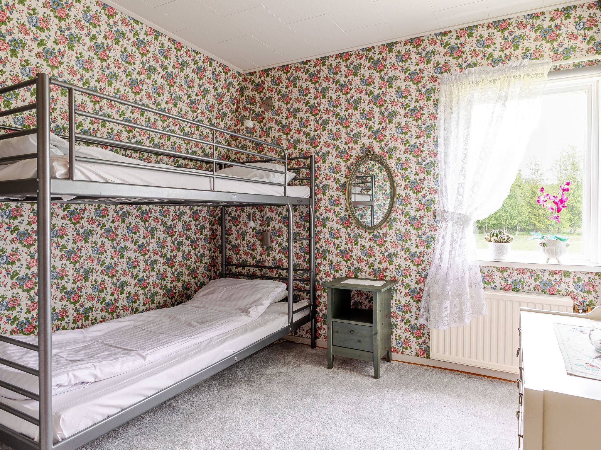 Une chambre avec un lit superposé, un papier peint à motifs floraux, une petite table, un miroir et une fenêtre donnant sur un paysage verdoyant.