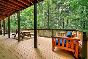 Terraza de madera con mesas de picnic y un banco con vistas a un bosque.