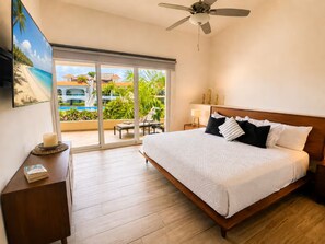 A hotel room with a bed, a nightstand, a bedside table, a ceiling fan, and a view of a pool and buildings outside. - Lagoon Front Infinity Pool Paddleboard And Fun Puerto Aventuras (Puerto Aventuras)