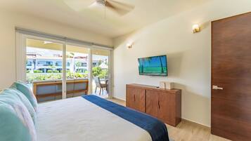 Un dormitorio con una cama grande, un armario de madera, un televisor y un ventanal corredero que da a un balcón con vistas a edificios y vegetación.