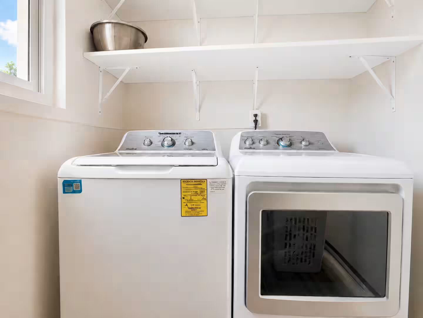 A laundry area with a front-loading washer and dryer, a metal shelf above, and a window to the left.