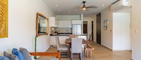 Una sala de estar moderna con un sofá, una mesa de comedor y sillas. Se ve un área de cocina al fondo con armarios y un lavamanos.