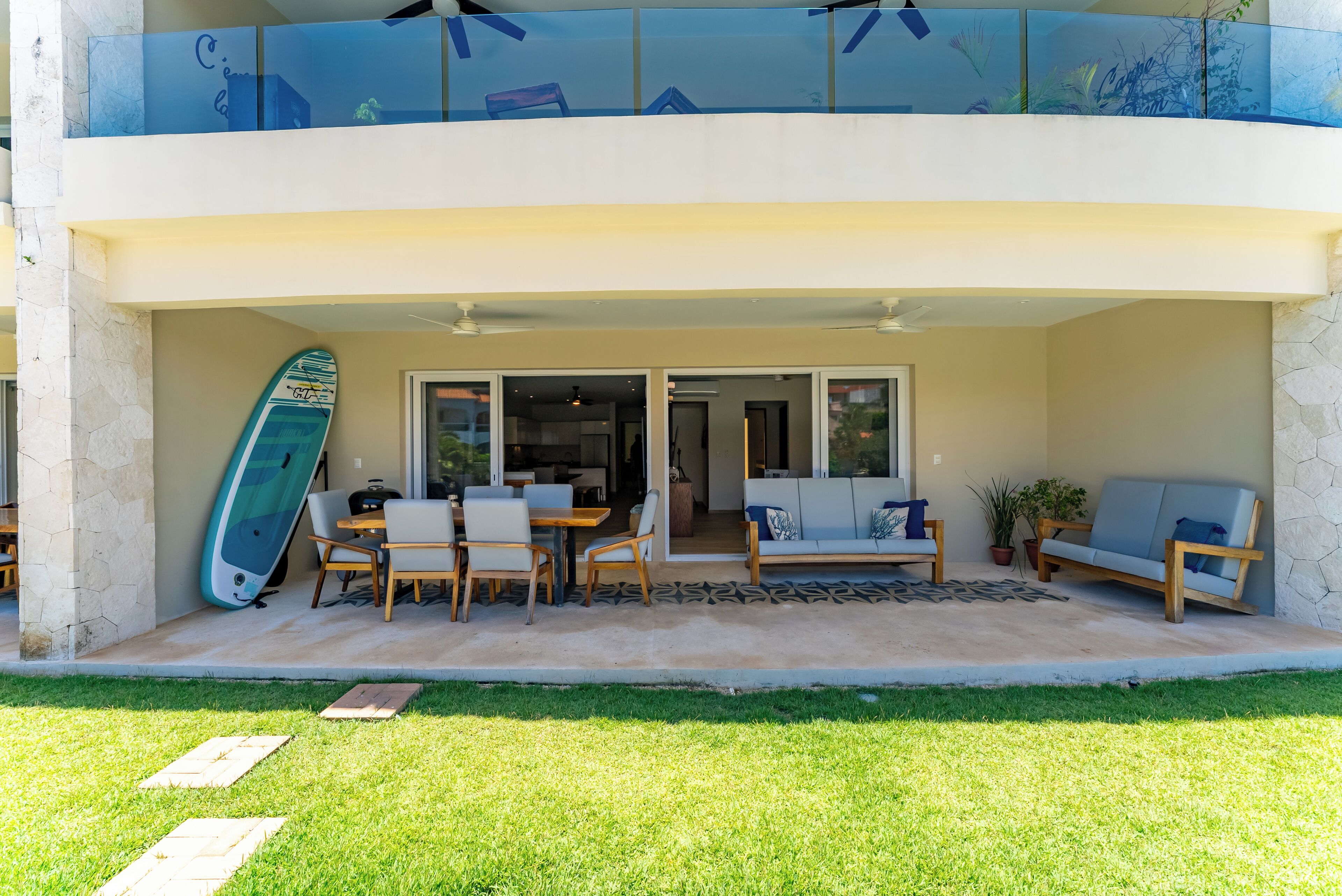 A two-story building with a covered patio area featuring outdoor furniture, a surfboard, and a glass balustrade.