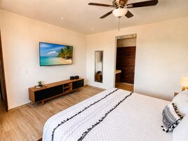 Un dormitorio con una cama, ventilador de techo, televisor, aparador de madera y una puerta que da a un baño.