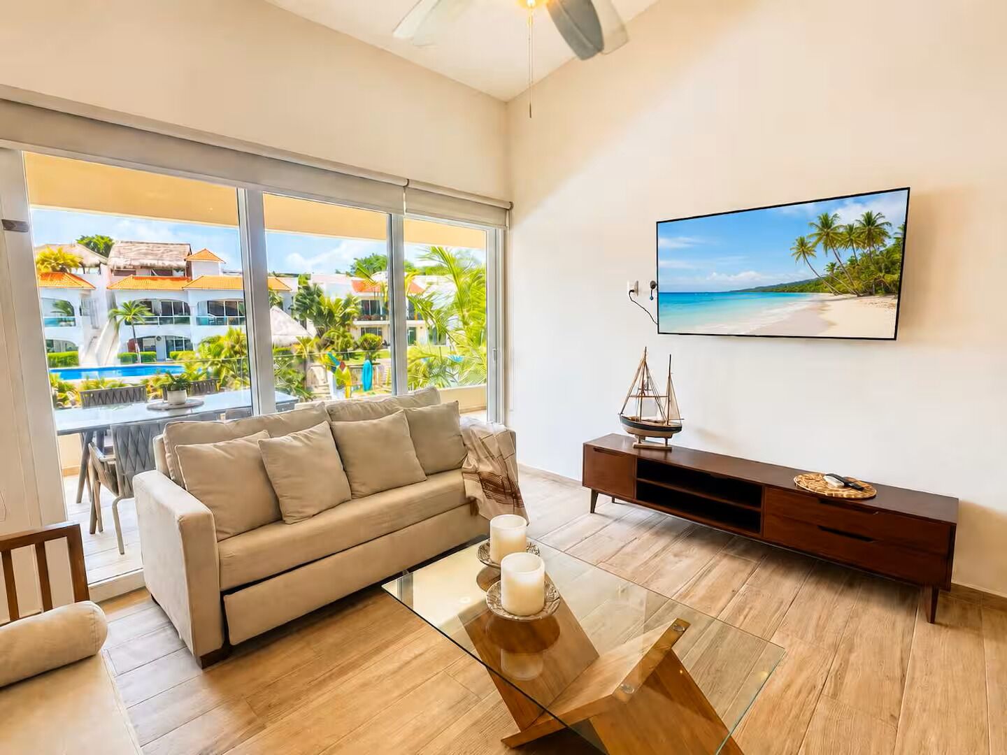 A modern living room with a sofa, a glass coffee table, a TV, and a view of a pool and buildings.