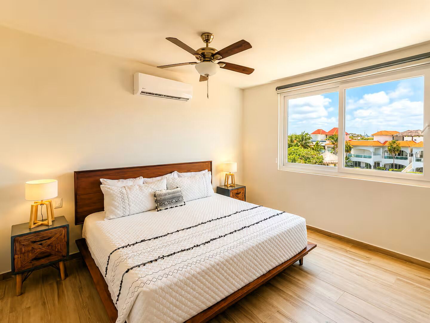 A bedroom with a bed, a ceiling fan, an air conditioner, bedside lamps, and a window with a view of buildings.