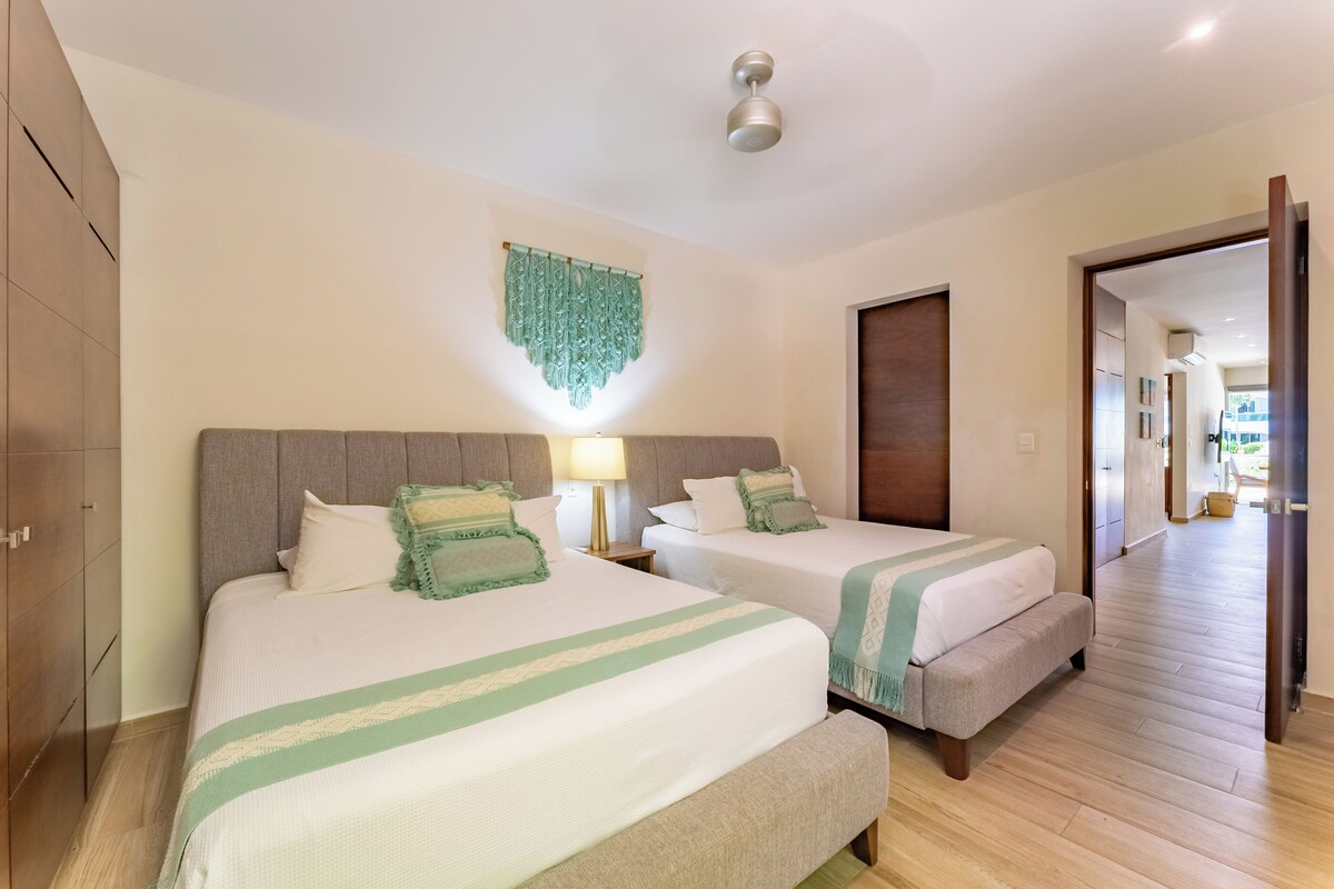 A hotel room with two beds, a wooden wardrobe, a ceiling fan, and a hallway leading to another room.