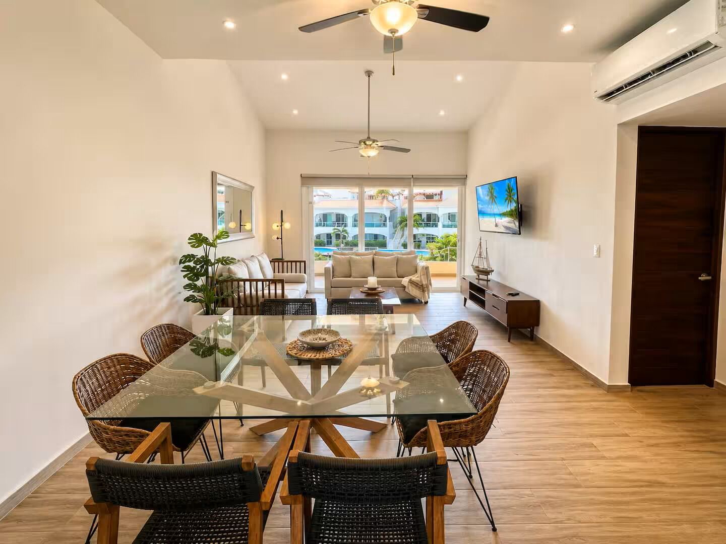 A spacious living area with a glass dining table, wicker chairs, a sofa, and a television mounted on the wall.