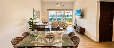 A spacious living area with a glass dining table, wicker chairs, a sofa, and a television mounted on the wall.