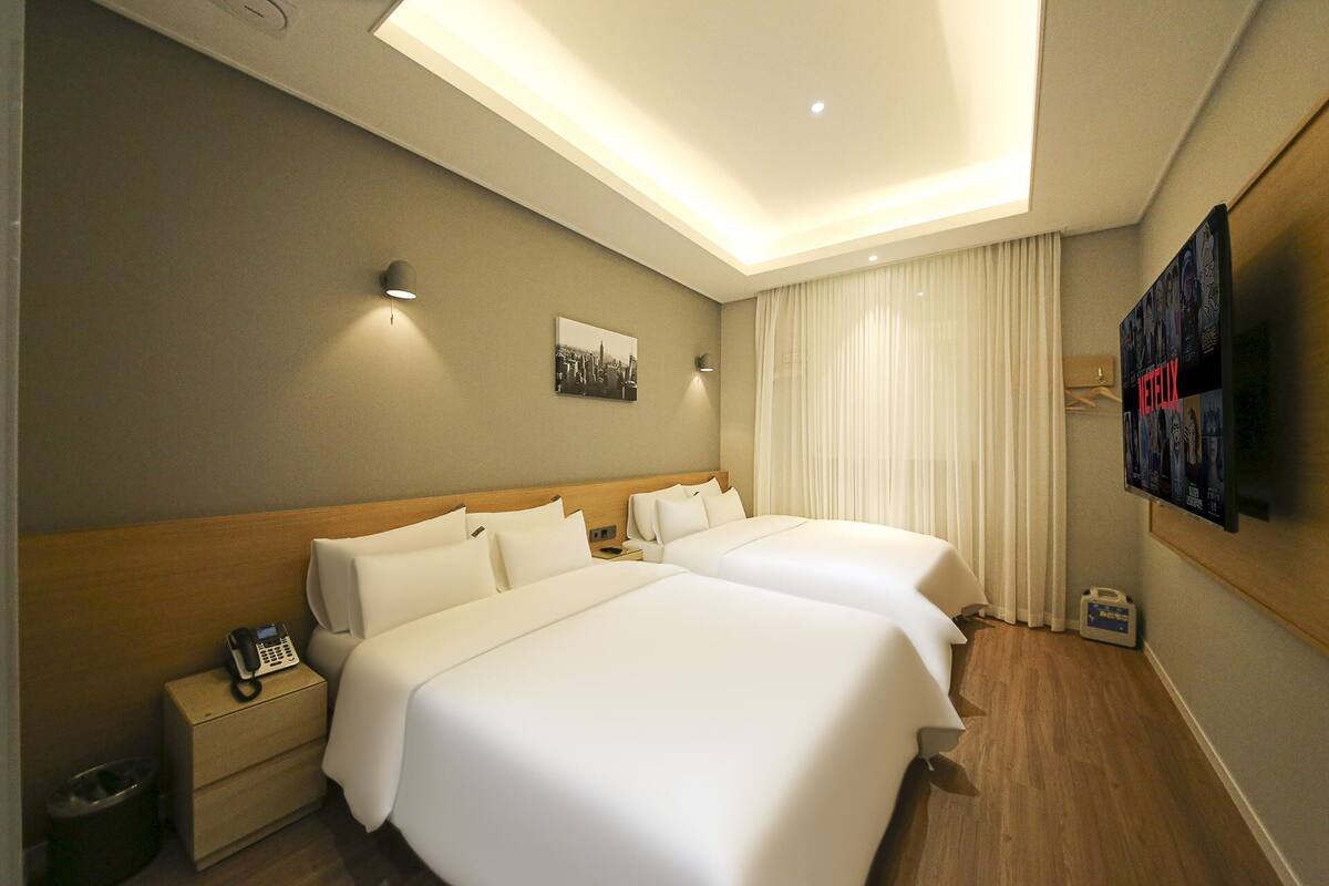 A hotel room with two beds, a bedside table with a phone, a trash bin, a wall-mounted TV, and a ceiling light.