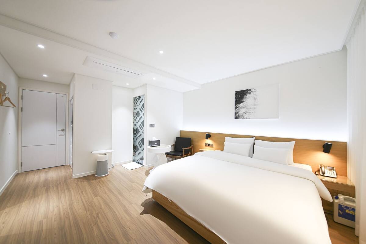 A modern hotel room with a large bed, a bedside table, a nightstand, a chair, a wardrobe, and a shower area.