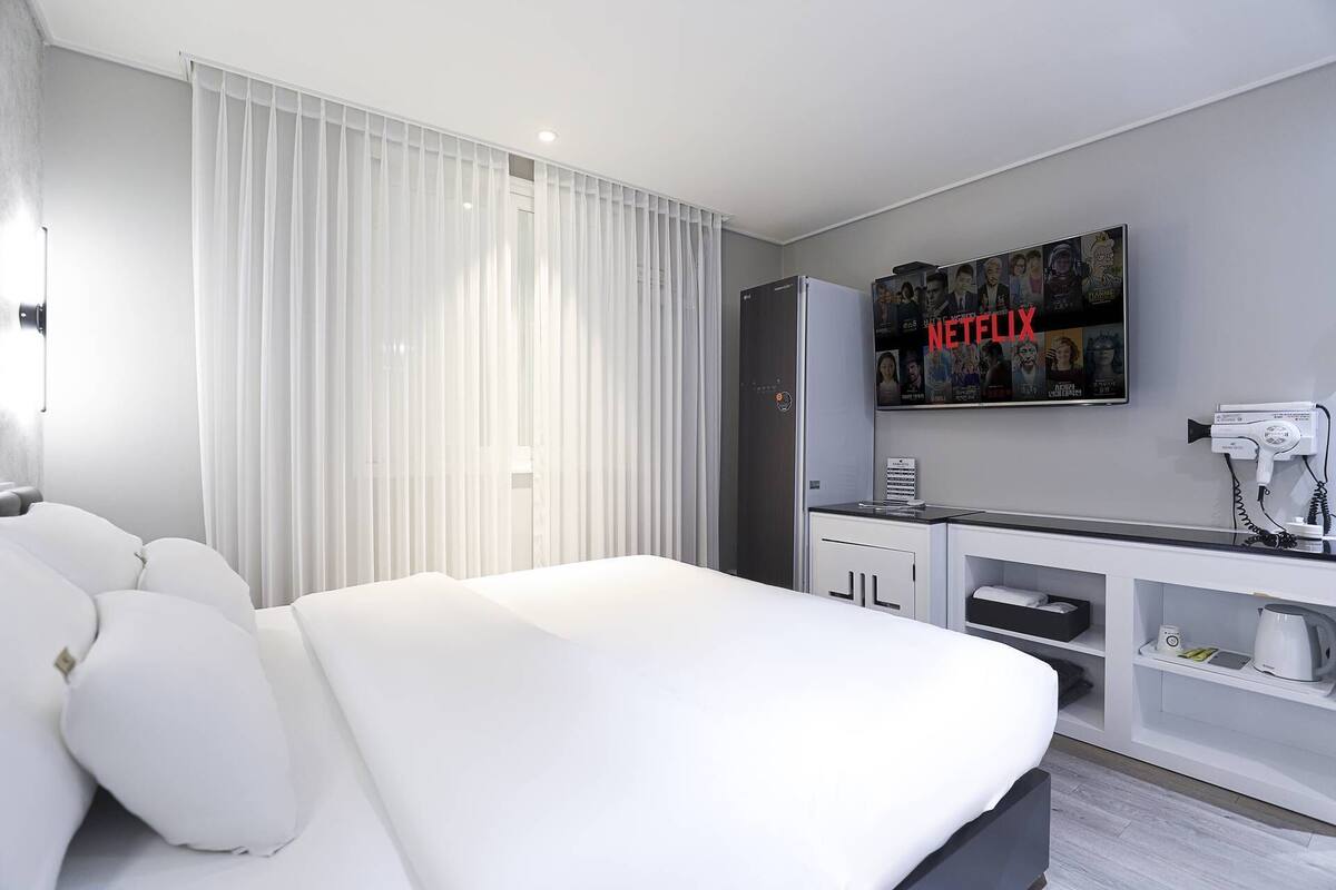 A modern hotel room with a large bed, a TV mounted on the wall displaying a Netflix logo, a minibar, and a hairdryer on the counter.