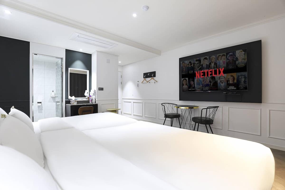 A hotel room with a large bed, a TV mounted on the wall displaying Netflix, a small table with chairs, and a bathroom visible through an open door.