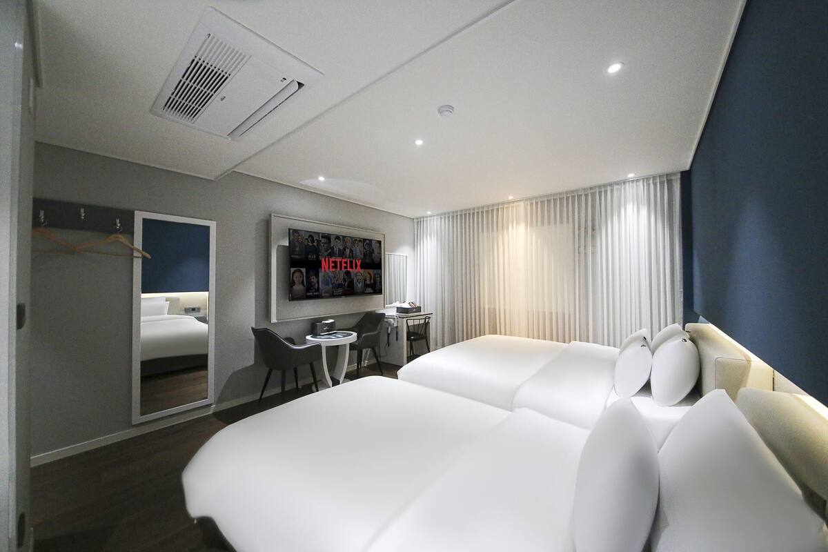 A modern hotel room with a large bed, a desk with a chair, a TV mounted on the wall, and a window with curtains.