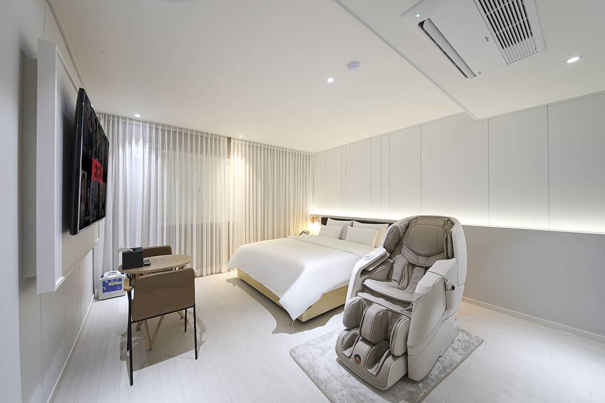 A modern hotel room with a large bed, a massage chair, a desk with a chair, and a TV mounted on the wall.