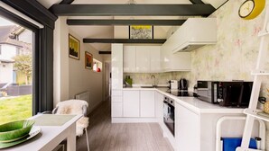 A modern kitchen with white cabinets, a built-in oven, and a microwave. There is a dining area with a white table and chairs, and a view of a garden outside. - Robin`s Nest, 1 bedroom near Wimbledon (London)