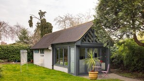 A small, single-story building with a sloped roof, large windows, and a garden area with a chair and a potted plant. - Robin`s Nest, 1 bedroom near Wimbledon (London)