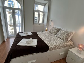 A bedroom with a bed, two bedside tables, a nightstand, and a window with a view of buildings.