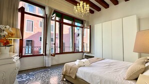 A bedroom with a large bed, a chandelier, and a view of buildings through open windows.